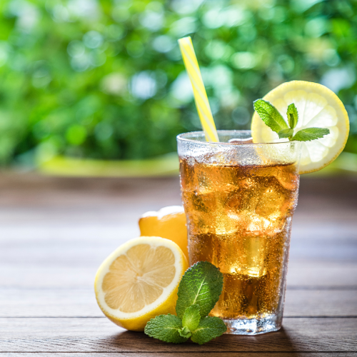 The image shows a refreshing iced tea with lemon slices and mint leaves on a wooden table, accompanied by a yellow straw, and a green, blurred background.