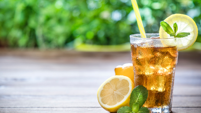 The image shows a refreshing iced tea with lemon slices and mint leaves on a wooden table, accompanied by a yellow straw, and a green, blurred background.