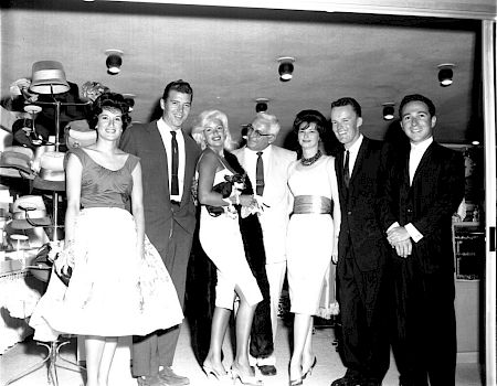 Seven people dressed formally are standing together and smiling for the camera in front of a display of hats in what appears to be a store.