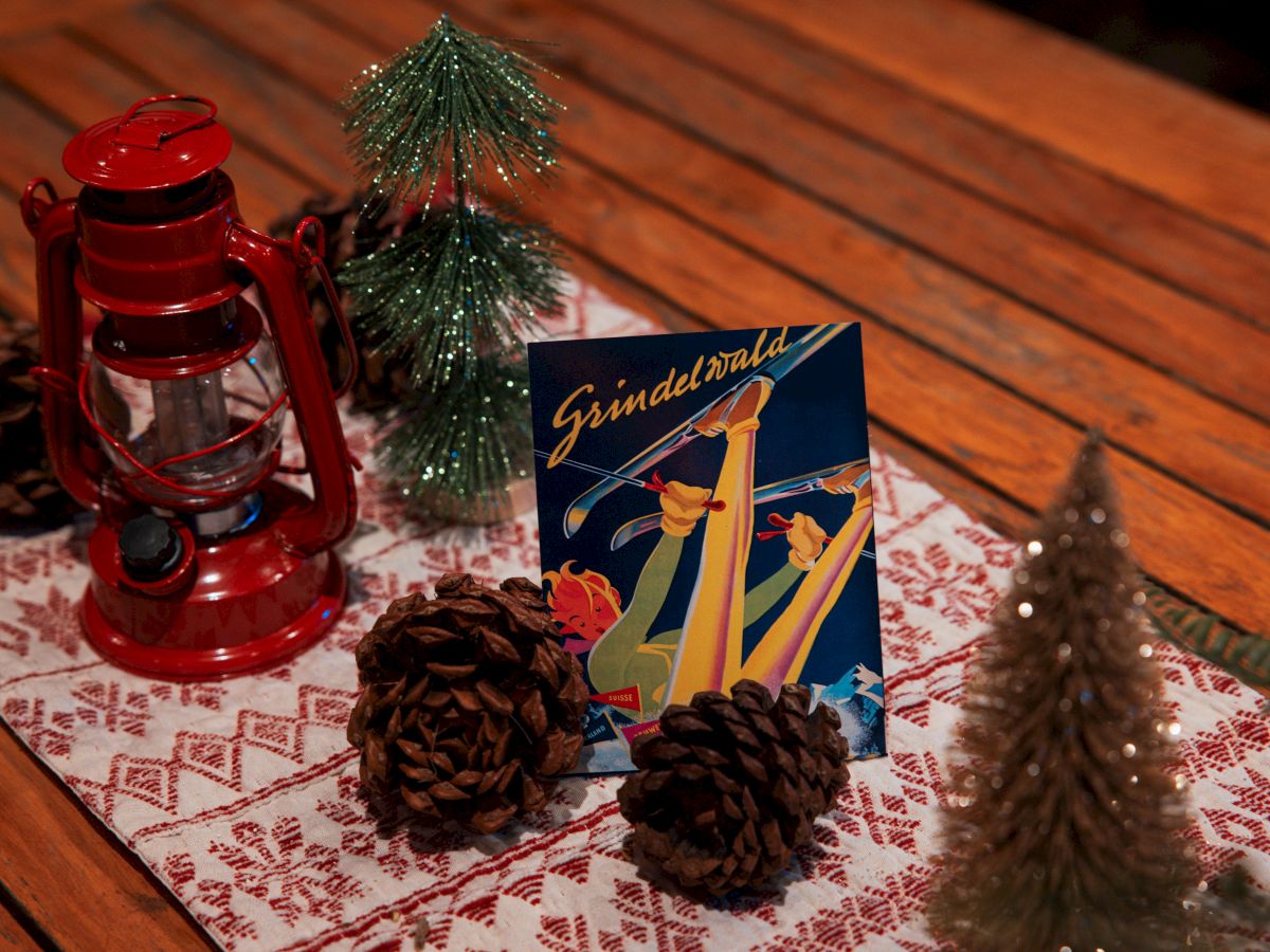 A holiday-themed setup with a red lantern, mini Christmas trees, pinecones, and a skiing-themed card on a wooden table with a festive runner.