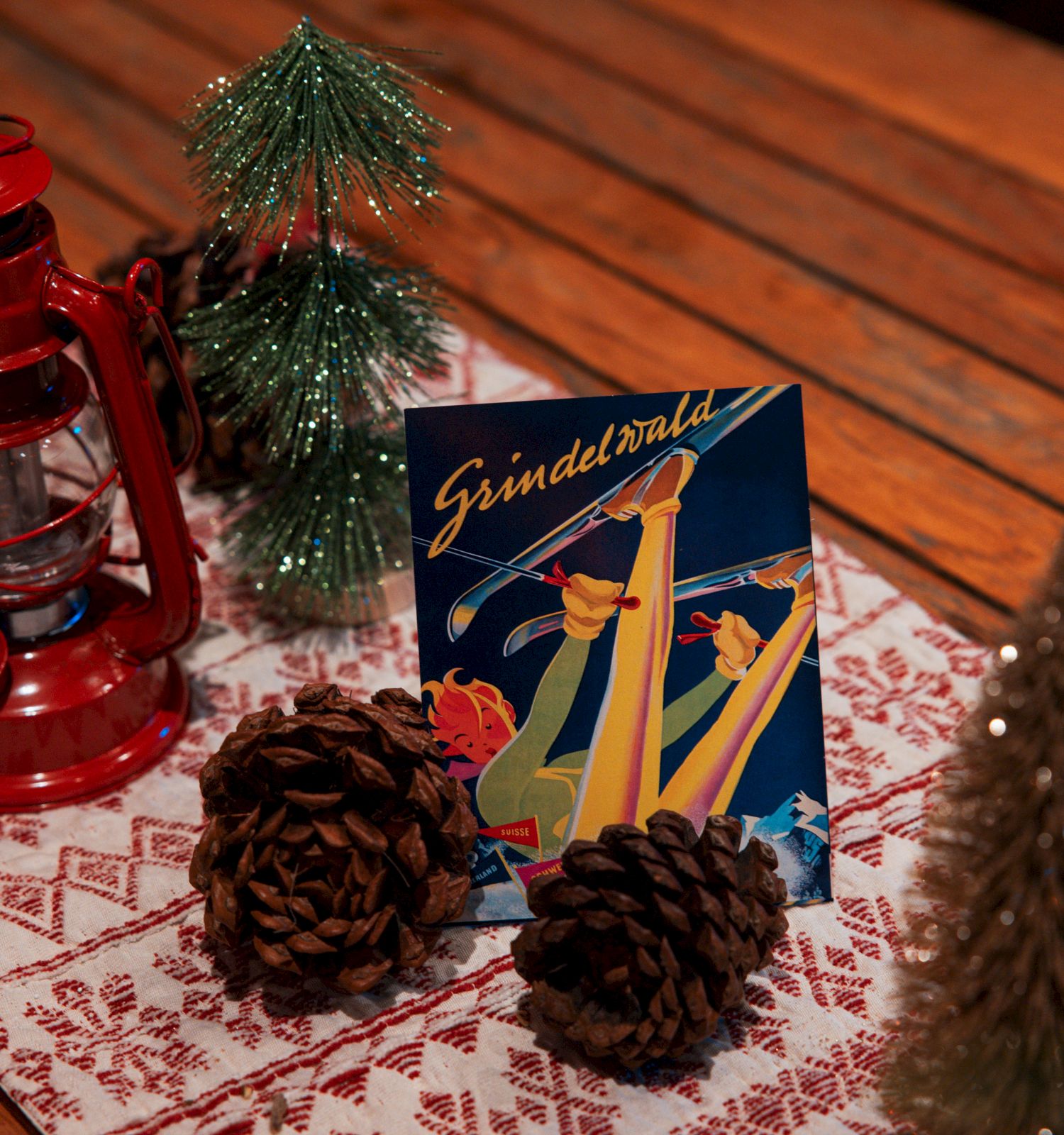 A holiday-themed setup with a red lantern, mini Christmas trees, pinecones, and a skiing-themed card on a wooden table with a festive runner.