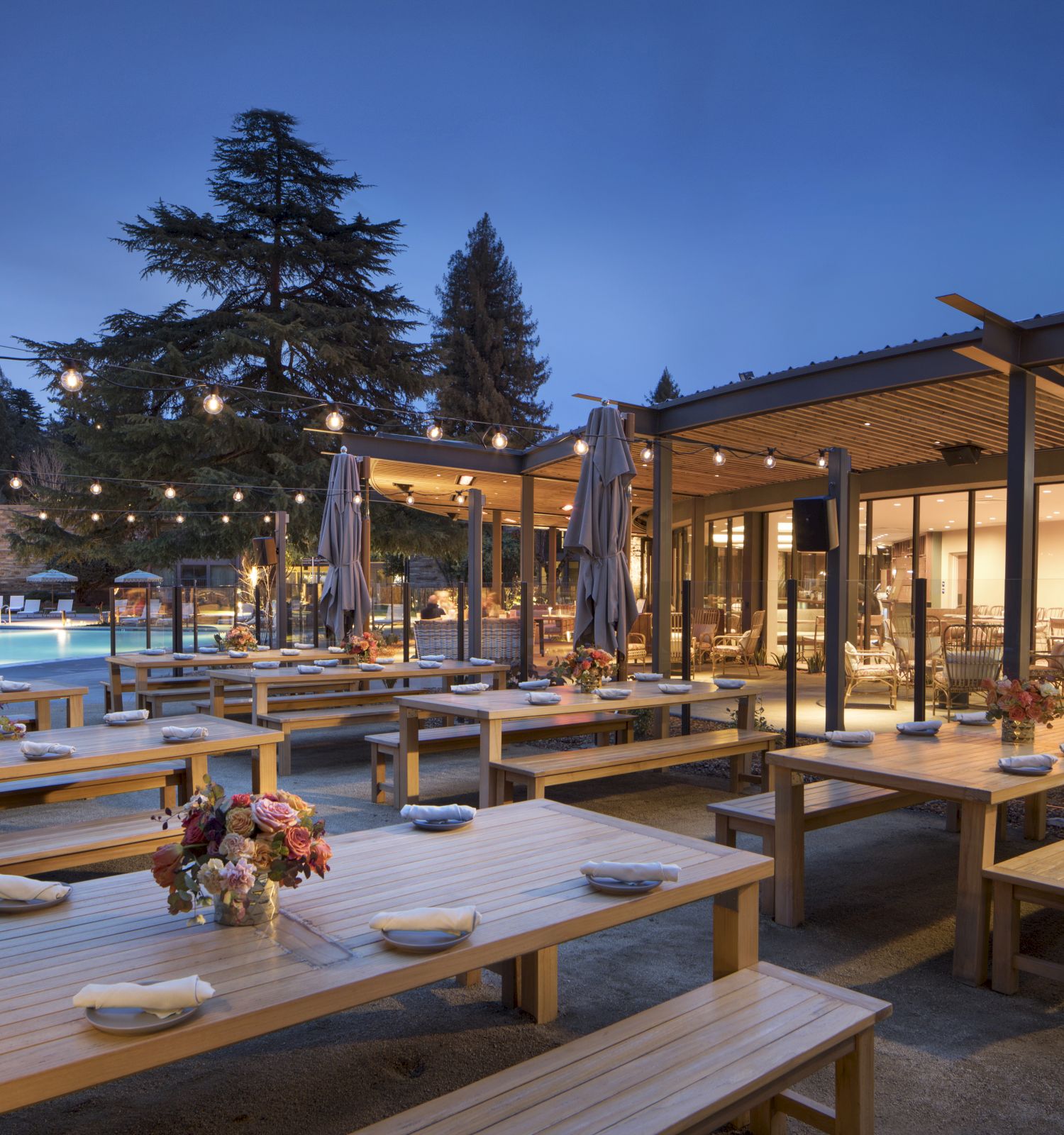 An outdoor dining area with wooden tables, benches, string lights, and floral centerpieces, near a building with large windows and a view of trees.