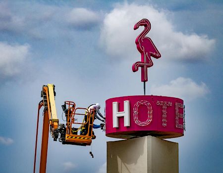 A worker on a lift is repairing a pink neon hotel sign featuring a flamingo against a backdrop of a partly cloudy sky.