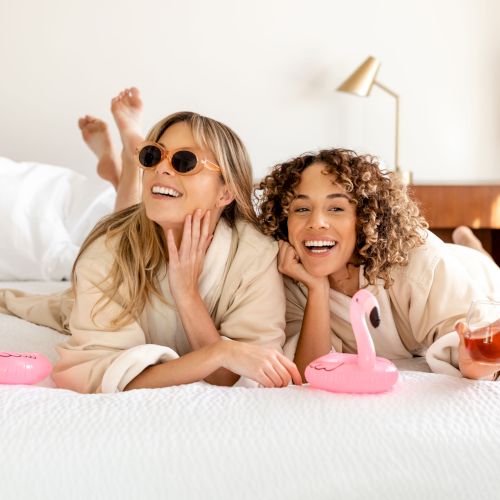 Two women in bathrobes relax on a bed with pink flamingo pool floats, one wearing sunglasses, both smiling, and one holding a beverage.