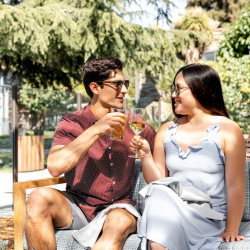 A man and a woman sit outdoors, toasting with drinks and smiling at each other, surrounded by food on a table and greenery in the background.