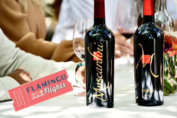 The image shows two wine bottles labeled “Muscardini” and “M,” alongside wine glasses on a table, plus a "Flamingo Flights" ticket.