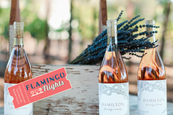 Three wine bottles labeled "HAMILTON" are displayed with a wooden box and flowers, featuring a "Flamingo Flights" tag beside them.