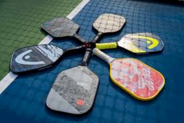 The image shows six pickleball paddles arranged in a star shape on a blue and green court surface.