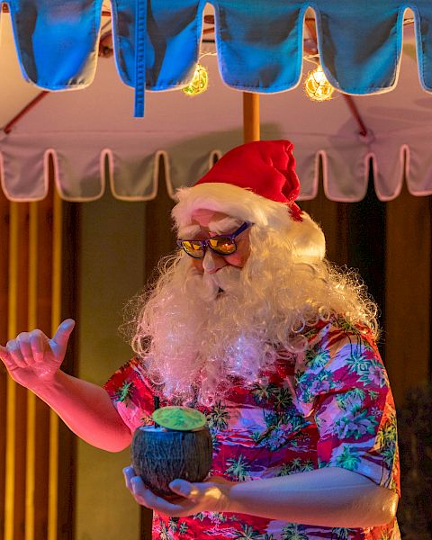 A person dressed as tropical-themed Santa with a red hat and floral shirt holds a drink under an outdoor umbrella, near a lit Christmas tree.