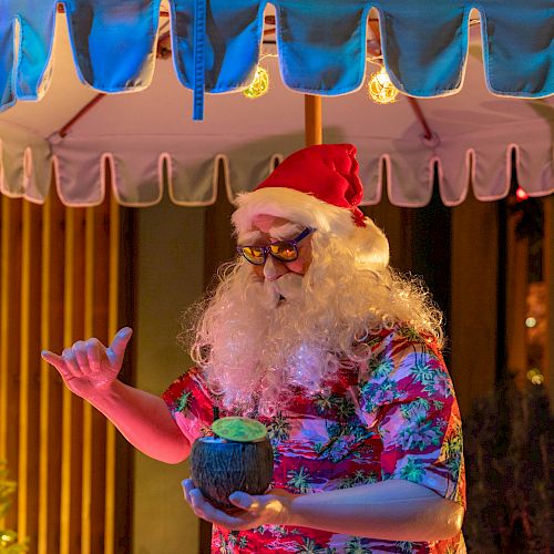 A person dressed as tropical-themed Santa with a red hat and floral shirt holds a drink under an outdoor umbrella, near a lit Christmas tree.