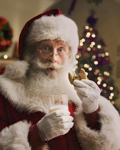 Santa Clause holding a glass of milk and a cookie, with a decorated Christmas tree in the background.