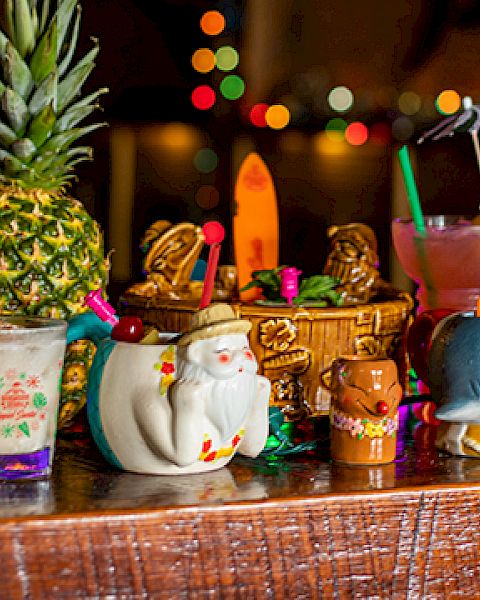 A collection of colorful tropical cocktails in themed mugs on a wooden bar, with festive lights in the background.