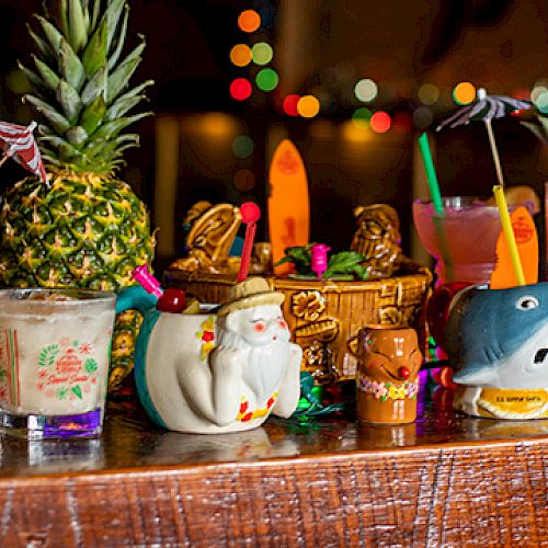 A collection of colorful tropical cocktails in themed mugs on a wooden bar, with festive lights in the background.