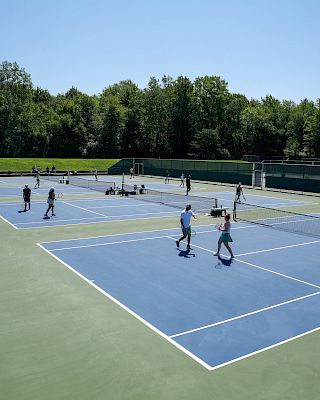 The image shows an outdoor tennis facility with multiple courts and people playing tennis under a clear sky, surrounded by trees and greenery.