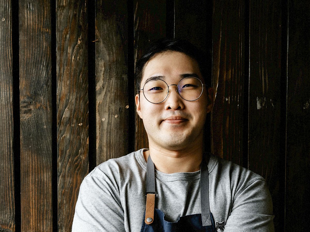 A person wearing a grey shirt and blue apron stands with arms crossed in front of a wooden wall, smiling at the camera.