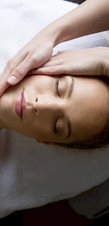 A person is lying down with eyes closed, receiving a relaxing face massage from another pair of hands, on a white towel.