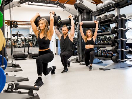 Three people in workout attire perform lunges with weights held overhead in a gym, surrounded by various fitness equipment.