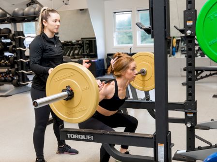 A person is performing a squat with a barbell while another person stands nearby in a gym setting, offering support or instruction.