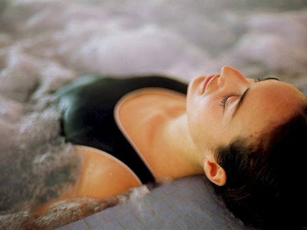 A person is relaxing in water, possibly in a pool or hot tub, with eyes closed and head resting on the pool edge, wearing a black swimsuit.