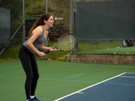 A woman is playing tennis, holding a racket and preparing for a shot on an outdoor tennis court.