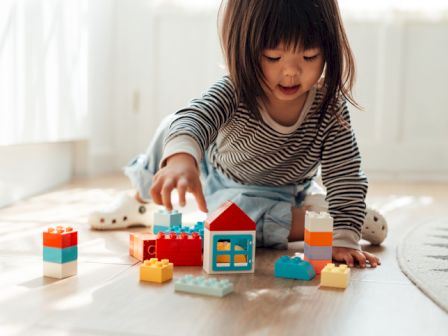 A child is sitting on the floor, playing with colorful toy building blocks and a small house in a bright room with wooden flooring.