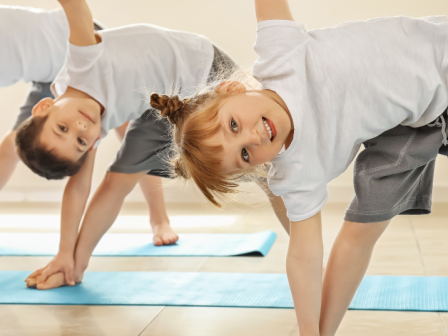 Children are practicing yoga, smiling, and spreading arms while standing on blue mats in a bright room.