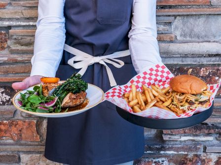 The image shows a server holding two dishes: one with a burger and fries, the other with steak, vegetables, and greens.