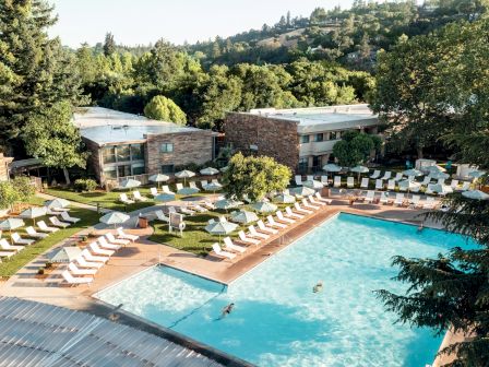 An outdoor pool area with numerous lounge chairs and umbrellas, surrounded by greenery and buildings, with a couple of people in the pool.