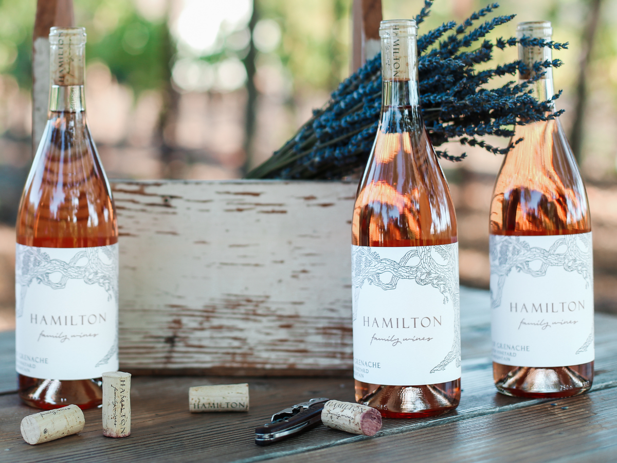 Three bottles of Hamilton wine, some corks, a wine opener, and a rustic wooden box with lavender flowers are arranged on a table.