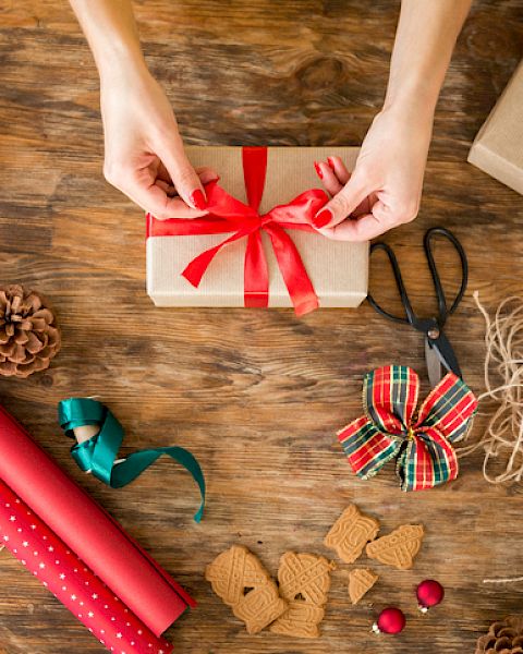 A person is tying a red ribbon around a wrapped gift on a wooden table, surrounded by wrapping paper, scissors, twine, and other festive items.