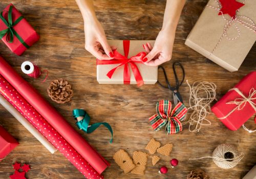 A person is tying a red ribbon around a wrapped gift on a wooden table, surrounded by wrapping paper, scissors, twine, and other festive items.