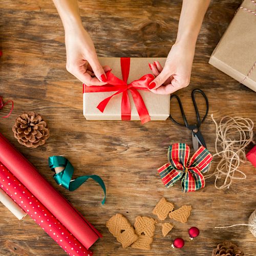 A person is tying a red ribbon around a wrapped gift on a wooden table, surrounded by wrapping paper, scissors, twine, and other festive items.