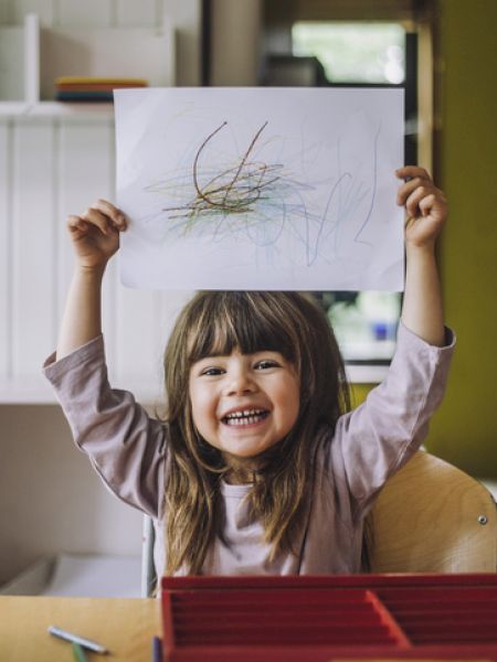 A child holds up a drawing with a big smile, sitting at a table with art supplies and a colorful scribble on the paper.