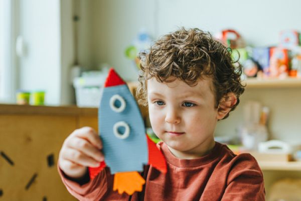 A child is playing with a handmade paper rocket in a colorful room with toys and decorations.