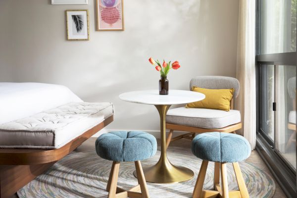 A cozy room with a bed, two blue stools, a white round table with tulips, and a chair by a window. Artwork is hung on the wall.