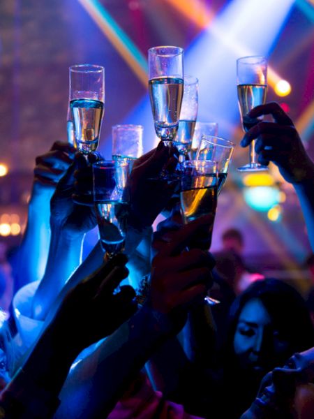 People raising champagne glasses in a celebratory toast under colorful, vibrant lighting at a party or festive gathering.