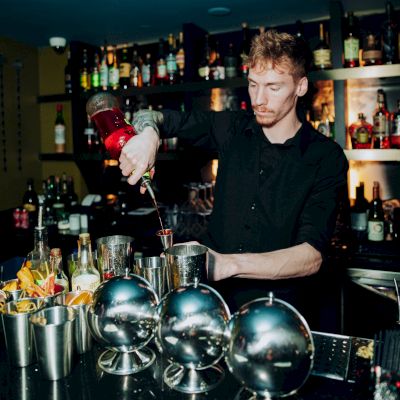 A bartender is mixing a drink behind a bar, surrounded by various bottles and utensils, in a dimly lit setting.