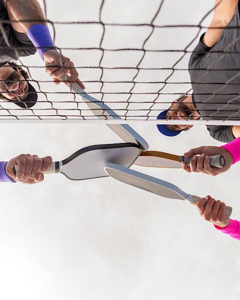 Four people near a tennis net holding badminton rackets towards the center, wearing colorful wristbands.