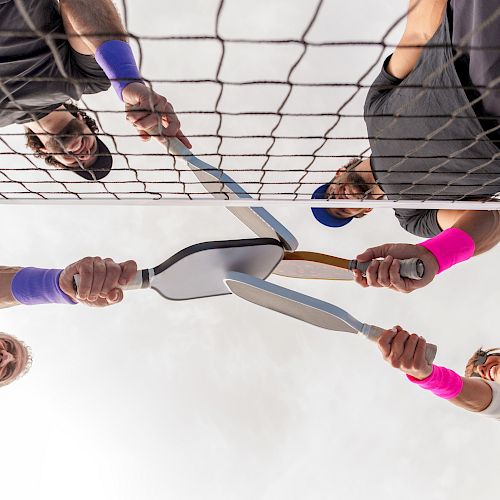 Four people near a tennis net holding badminton rackets towards the center, wearing colorful wristbands.