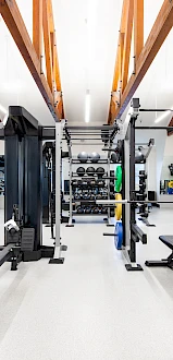 A well-lit gym with wooden beams features treadmills, weight benches, fitness machines, and free weights with mirrors lining the walls.