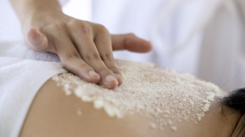 A person is receiving a back massage with a scrub being applied by a hand. The focus is on the back and hand.