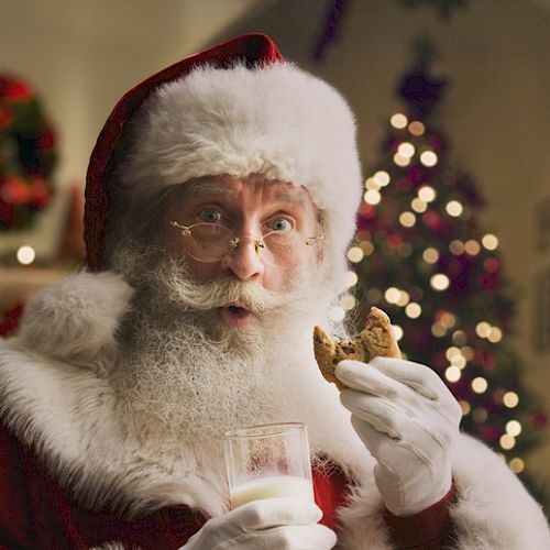 Santa Clause holding a glass of milk and a cookie, with a decorated Christmas tree in the background.