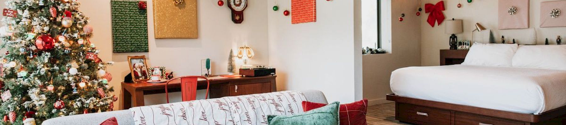 A festive room with a decorated Christmas tree, garlands, and a cozy couch with pillows. A bed is in the background, adding warmth.