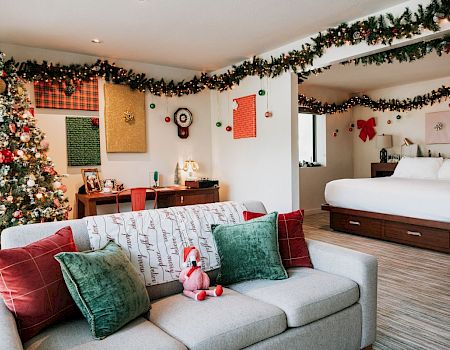 A festive room with a decorated Christmas tree, garlands, and a cozy couch with pillows. A bed is in the background, adding warmth.