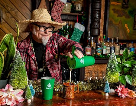 A person in a straw hat pours a drink in a festive setting, surrounded by plants, colorful trees, and flowers on a wooden bar.