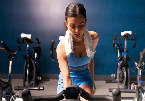 A person is on an exercise bike in a gym, wearing a blue outfit with a towel around their neck.