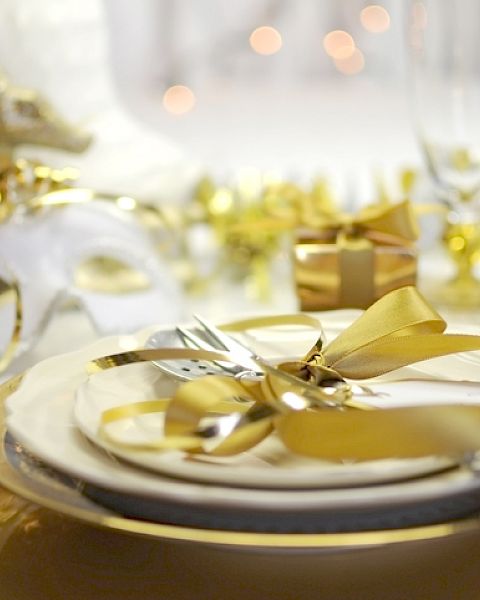 A festive table setting with gold accents, a mask, and a ribbon-wrapped gift against a blurred background with glowing lights.