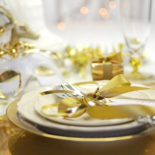 A festive table setting with gold accents, a mask, and a ribbon-wrapped gift against a blurred background with glowing lights.