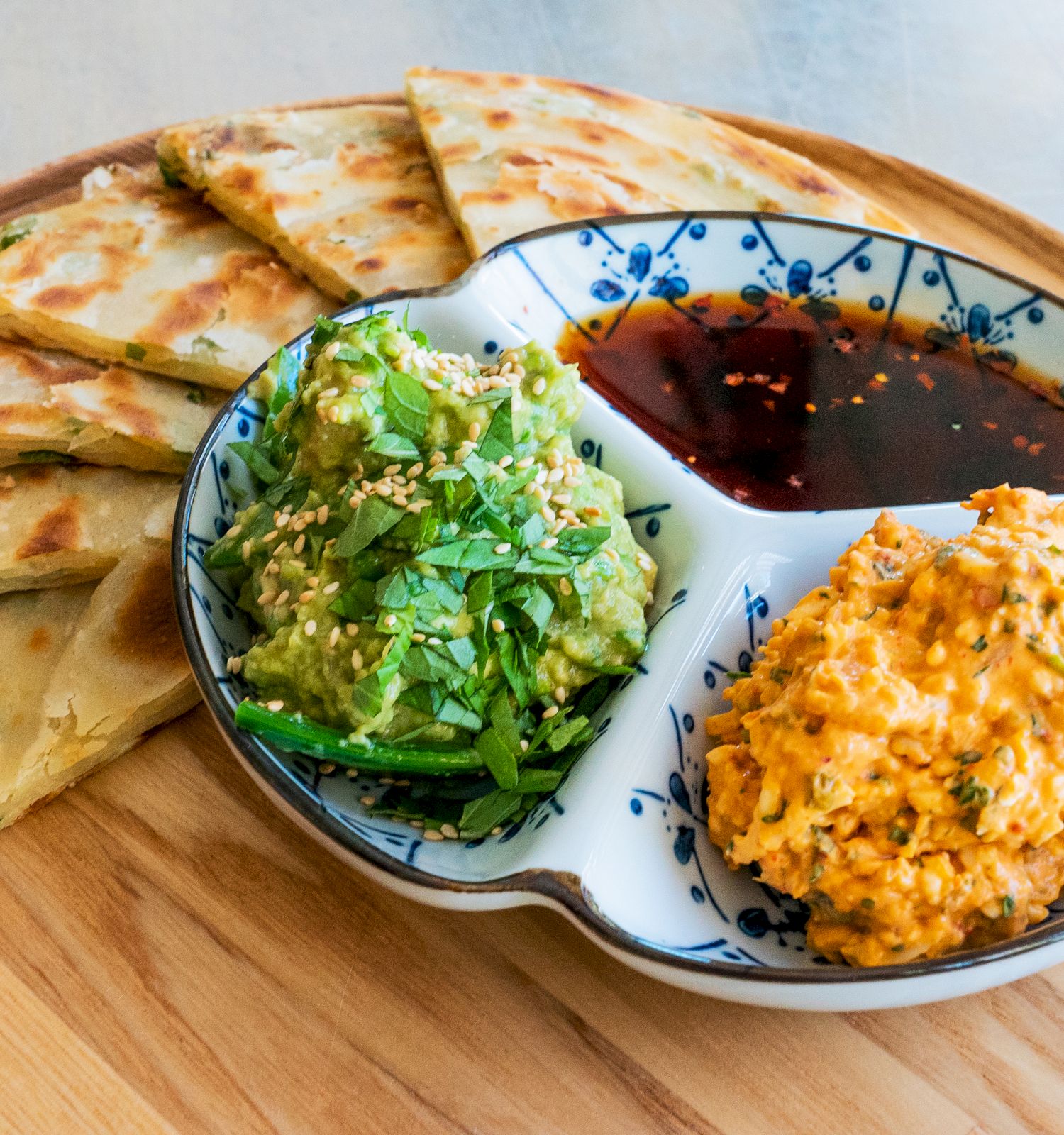 The image shows sliced scallion pancakes with dips: guacamole, a reddish sauce, and a carrot-like spread on a decorative plate.