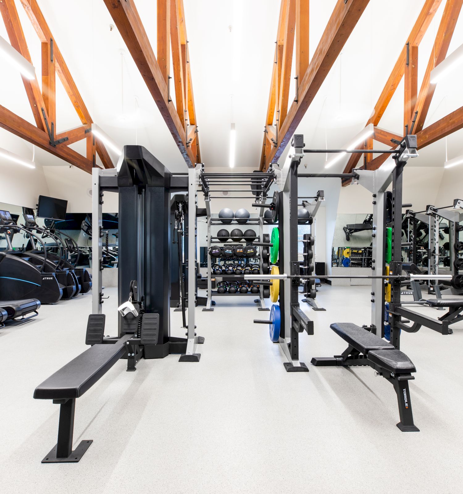 A modern gym with treadmills, weights, benches, and weightlifting racks, under wooden ceiling beams. Bright and organized for workouts.